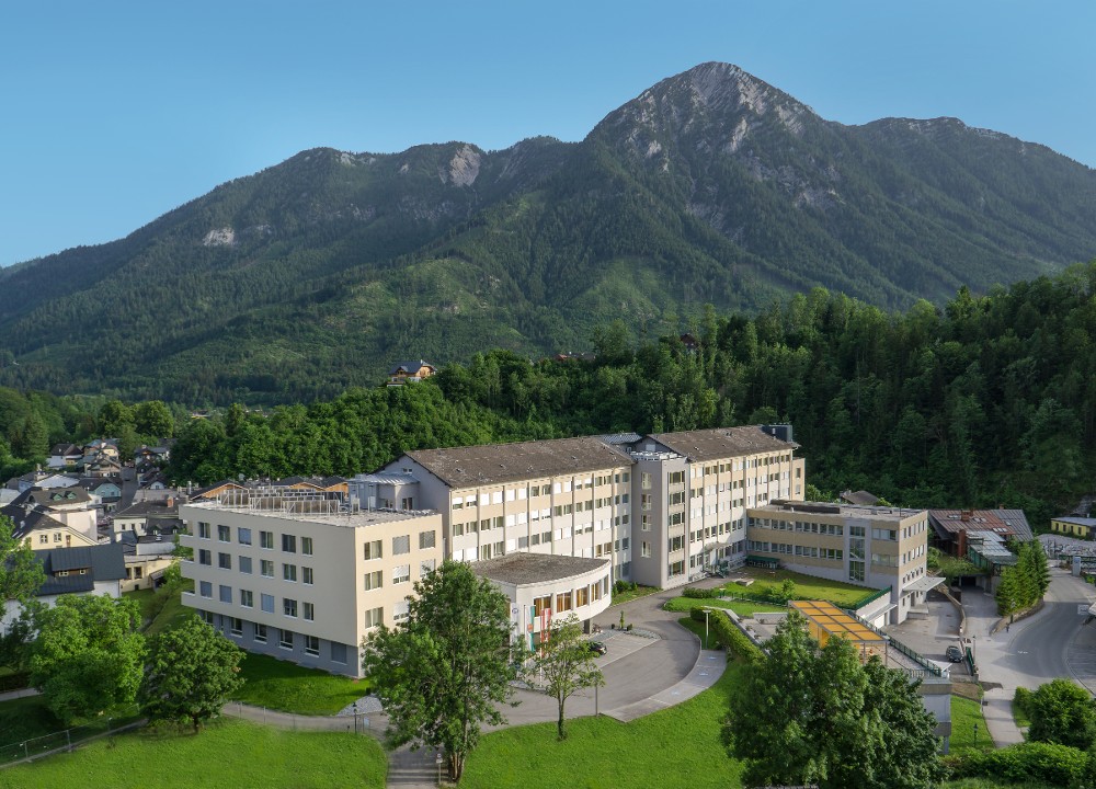Blick von oben auf das Reha-Zentrum Bad Aussee mit bewaldetem Hang und Bergmassiv im Hintergrund